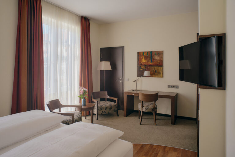 Studio-Hotelzimmer: zwei Betten, Schreibtisch, Sessel, TV, Fenster.