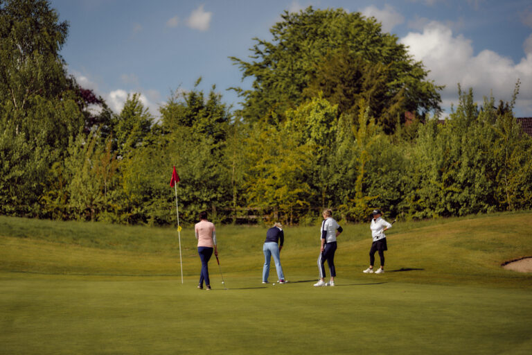 Vier Golfer auf dem Grün; einer puttet, während die anderen in der Nähe zusehen.