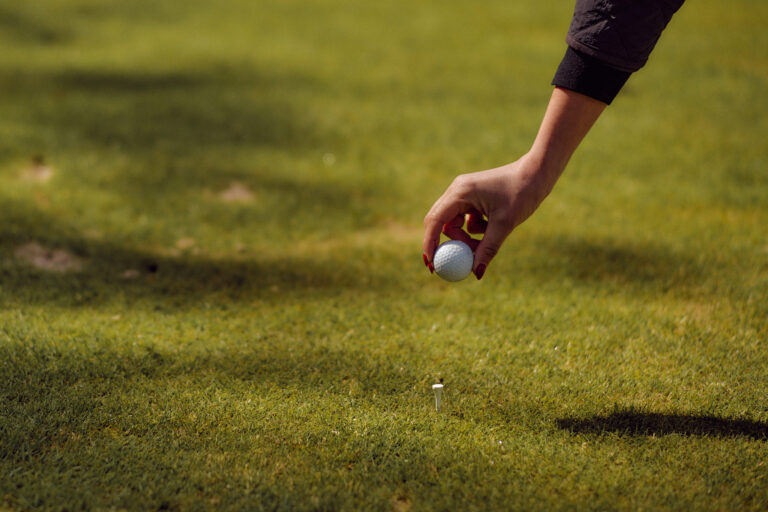 Eine rotgenagelte Hand legt den Golfball auf den Abschlag und wirft einen Schatten.