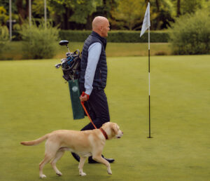 Ein Mann mit einer Golftasche führt einen gelben Labrador Retriever an einer roten Leine über den grünen Golfplatz des Golfclubs Treudelberg, vorbei an einer Fahnenstange mit Bäumen und Büschen im Hintergrund.