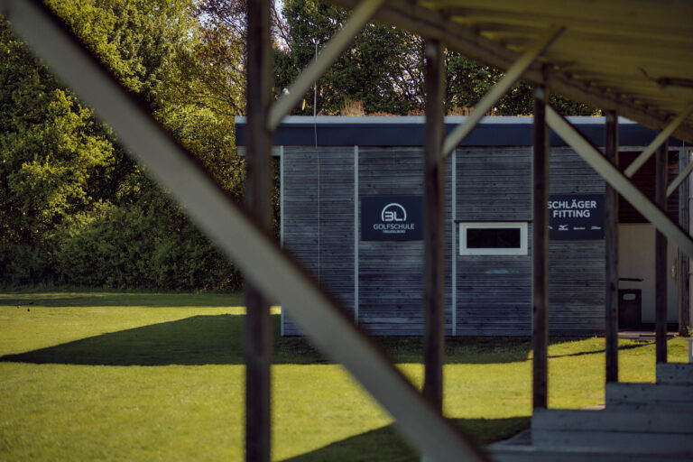 Hölzernes Gebäude der "Golfschule" inmitten von Grün, teilweise verdeckt durch ein Gebäude.