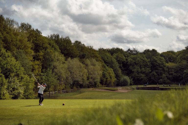 Golfer schwingt auf grünem Fairway in der Nähe von Bäumen und Hotel Hamburg.