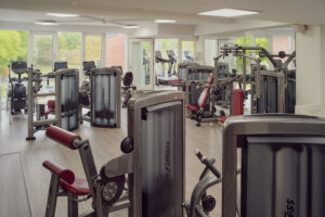 Moderner Fitnessraum mit verschiedenen Geräten, Holzböden, Gartenblick.