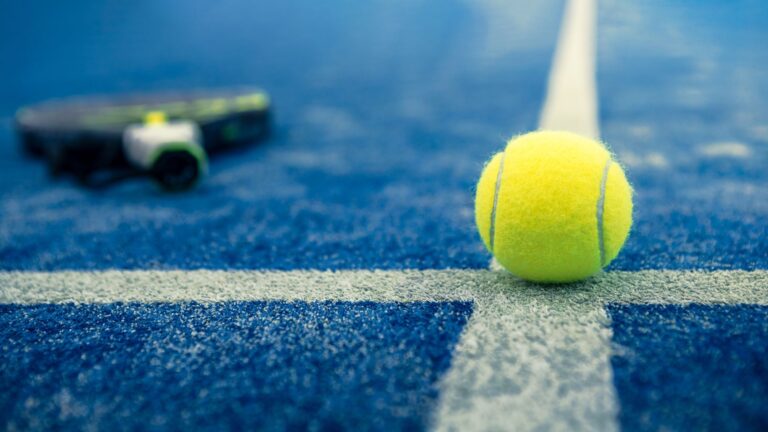 Gelber Tennisball auf blauem Platz, Schläger unscharf im Hintergrund.