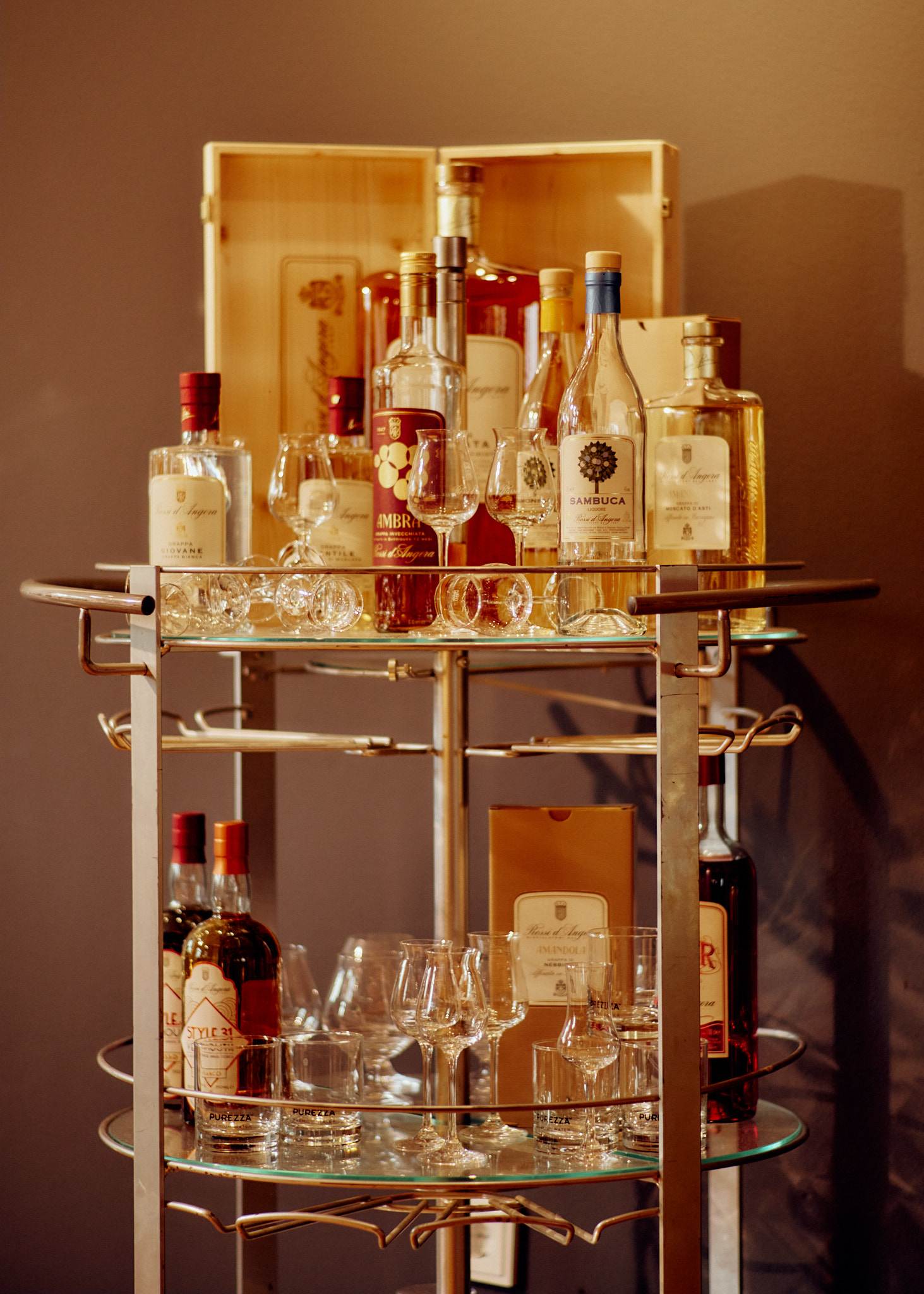 Pizzeria-noi-bar-cart zeigt einen Barwagen aus Metall mit Flaschen, Glaswaren, Holzkiste und braunem Wandhintergrund.