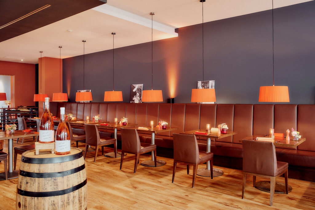 Das Interieur der Pizzeria_NOI_restaurant zeigt braune Ledersitze, Holztische, ein Weinfass in der Mitte und orangefarbene Hängelampen.