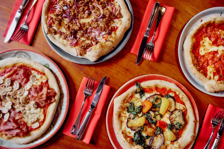 PizzeriaNOI_four-pizzas-woody-table zeigt vier Pizzen mit verschiedenen Belägen, jeweils mit Besteck und roten Servietten.