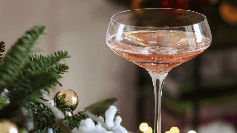 Christmas brunch coupe pink pearl wine glass next to snow-covered fir branch with golden ornaments for Christmas party.