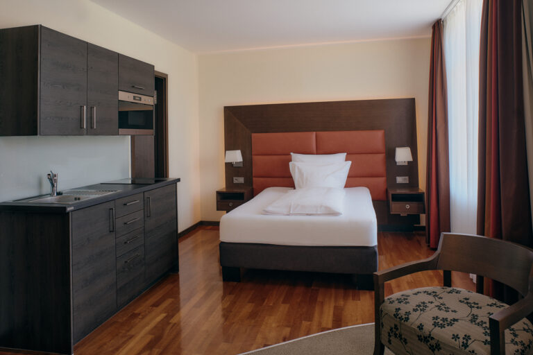 Das ModernBusinessZimmer verfügt über ein Einzelbett, eine Küchenzeile mit dunklen Schränken, einen gemusterten Stuhl und große Fenster mit Vorhängen.