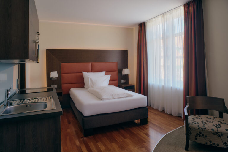 Business-Zimmer-Einzelzimmer-Hotelzimmer mit weißem Bettzeug, Kopfteil, Küchenzeile, Stuhl, Parkettboden und Fenster mit Vorhängen.