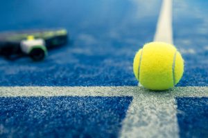 Gelber Tennisball auf blauem Platz, Schläger unscharf im Hintergrund.